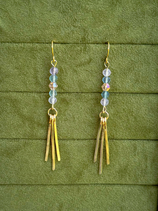 Handmade earrings with small iridescent beads and three gold-toned fringe bars, displayed on a textured olive green fabric background.