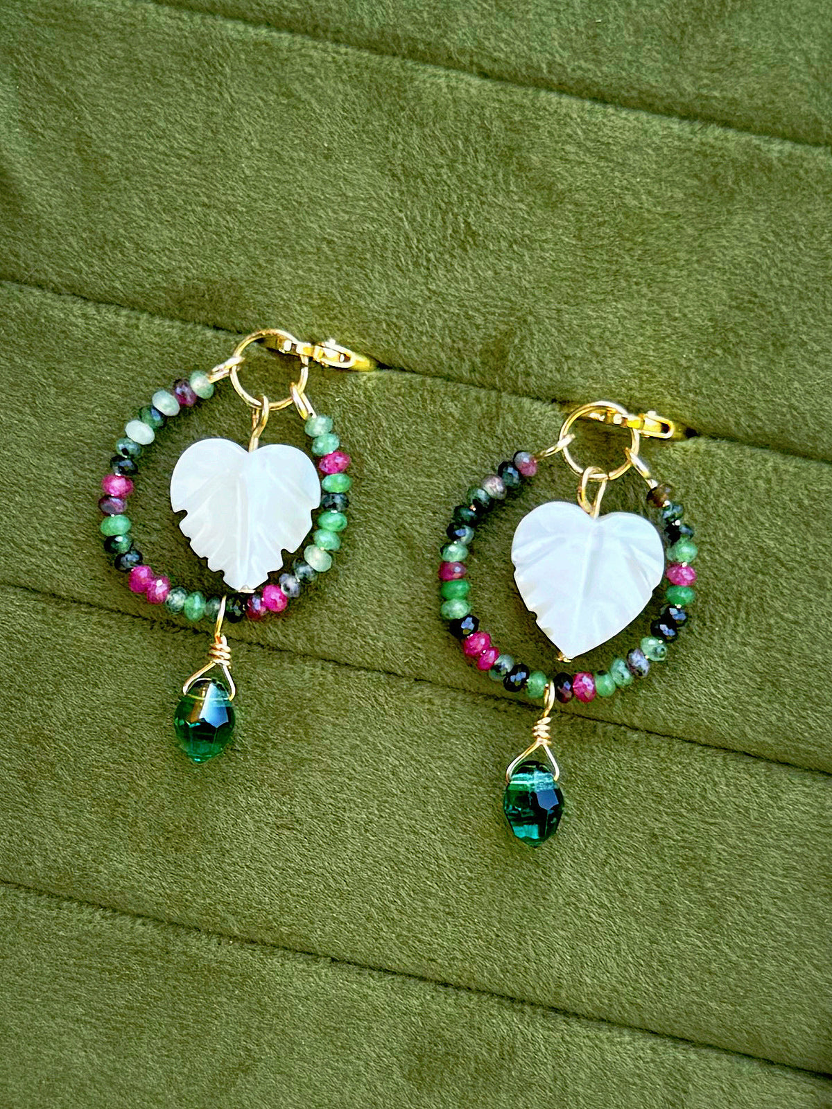 Handmade hoop earrings with gold wire, featuring a central white carved leaf charm, surrounded by small multicolored beads (green, pink, purple, black), and a dangling faceted green gemstone, displayed on an olive green textured background.