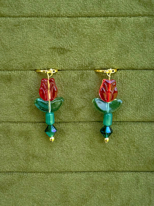 Handmade earrings with red flower-shaped beads, green leaves, and green faceted beads on gold huggies, displayed on a textured olive green background.