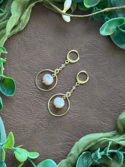 Handmade earrings with circular gold wire frames and freshwater pearl beads, arranged on a natural surface surrounded by green leaves.
