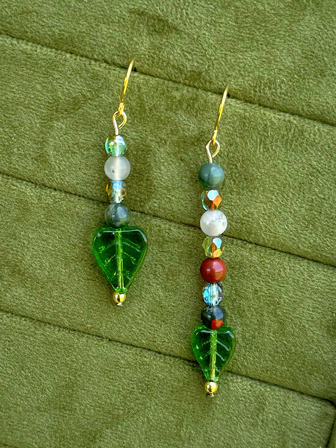 Handmade asymmetrical beaded earrings with gemstone and glass beads displayed on a textured olive green fabric background.