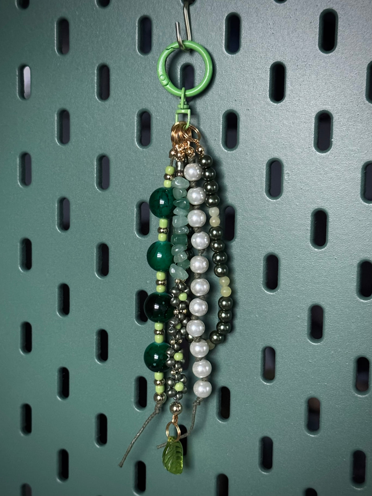 Handmade beaded keychain or bag charm with green and white beads, a gold clasp, and a green keyring, laid on a brown canvas surface.