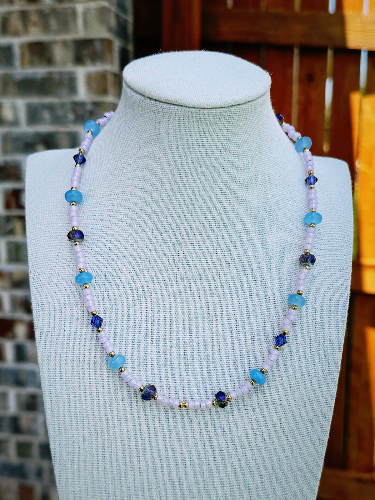 Handmade beaded necklace displayed on a fabric jewelry bust. The necklace features light purple lavender beads, light blue beads, dark blue faceted beads, and small gold spacer beads.