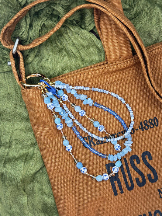 Handmade bag charm with beads of light blue, medium blue, and white. The bag is laid flat on a textured olive green fabric background.