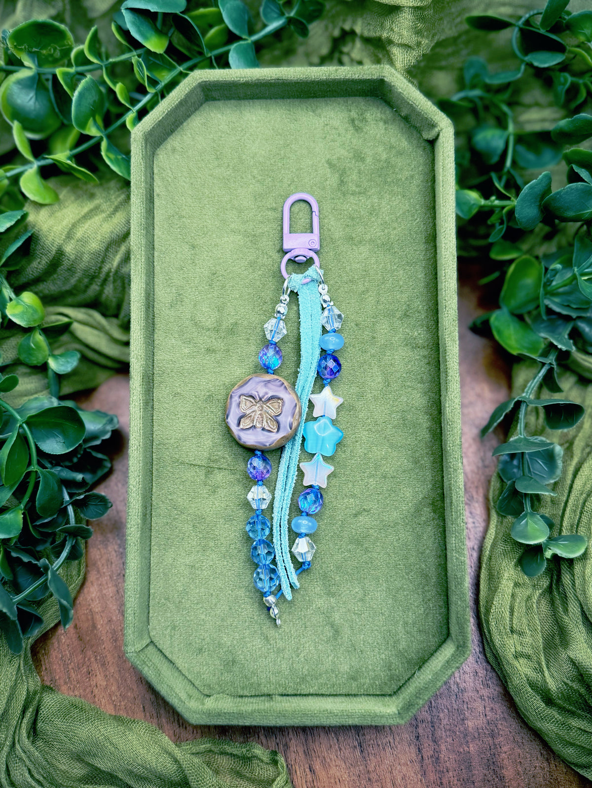 Handmade beaded charm keychain with a lavender clasp, light blue tassel, blue beads, and a butterfly charm, presented on an octagonal green velvet tray surrounded by green leaves.