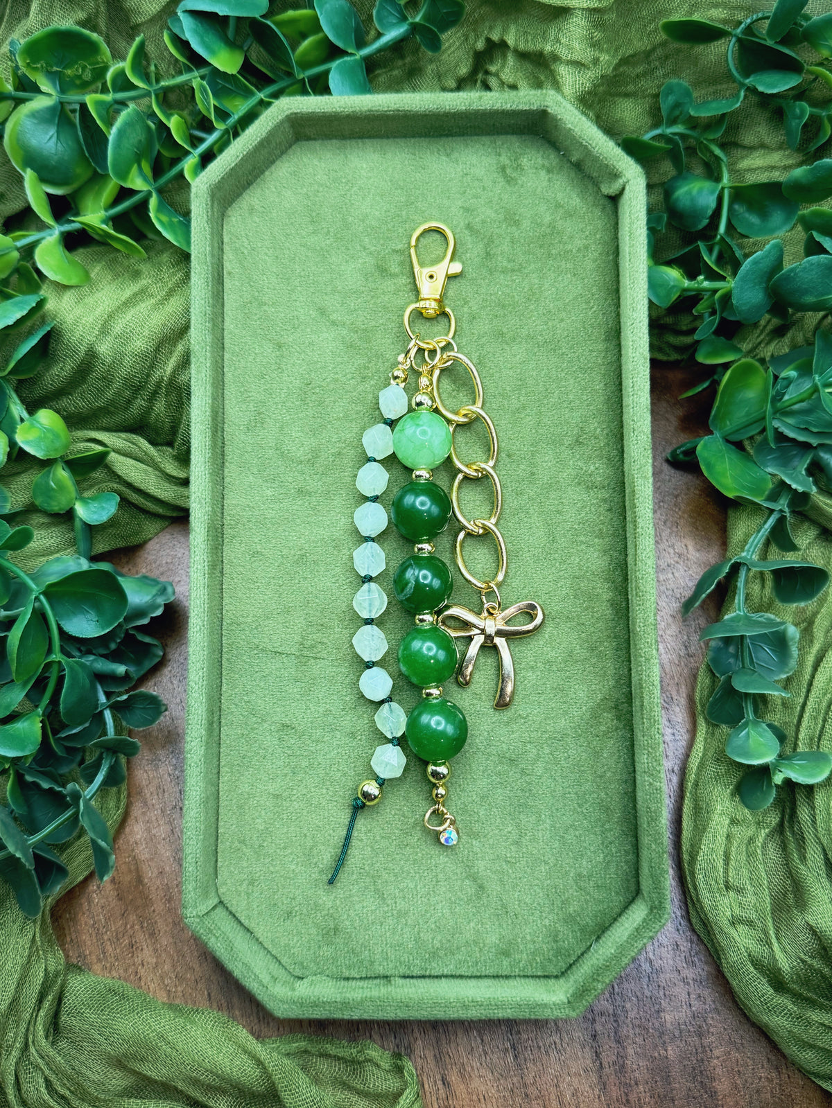 Handmade beaded keychain or bag charm with green and white beads, gold-tone rings, a lobster clasp, and a small dragonfly charm, displayed on a green velvet tray surrounded by green leaves and fabric.
