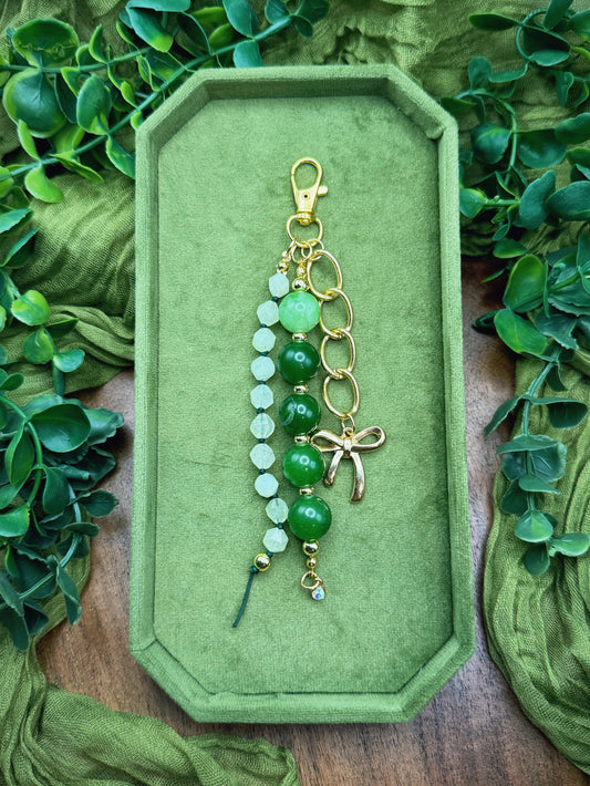Handmade beaded keychain or bag charm with green and white beads, gold-tone rings, a lobster clasp, and a small dragonfly charm, displayed on a green velvet tray surrounded by green leaves and fabric.
