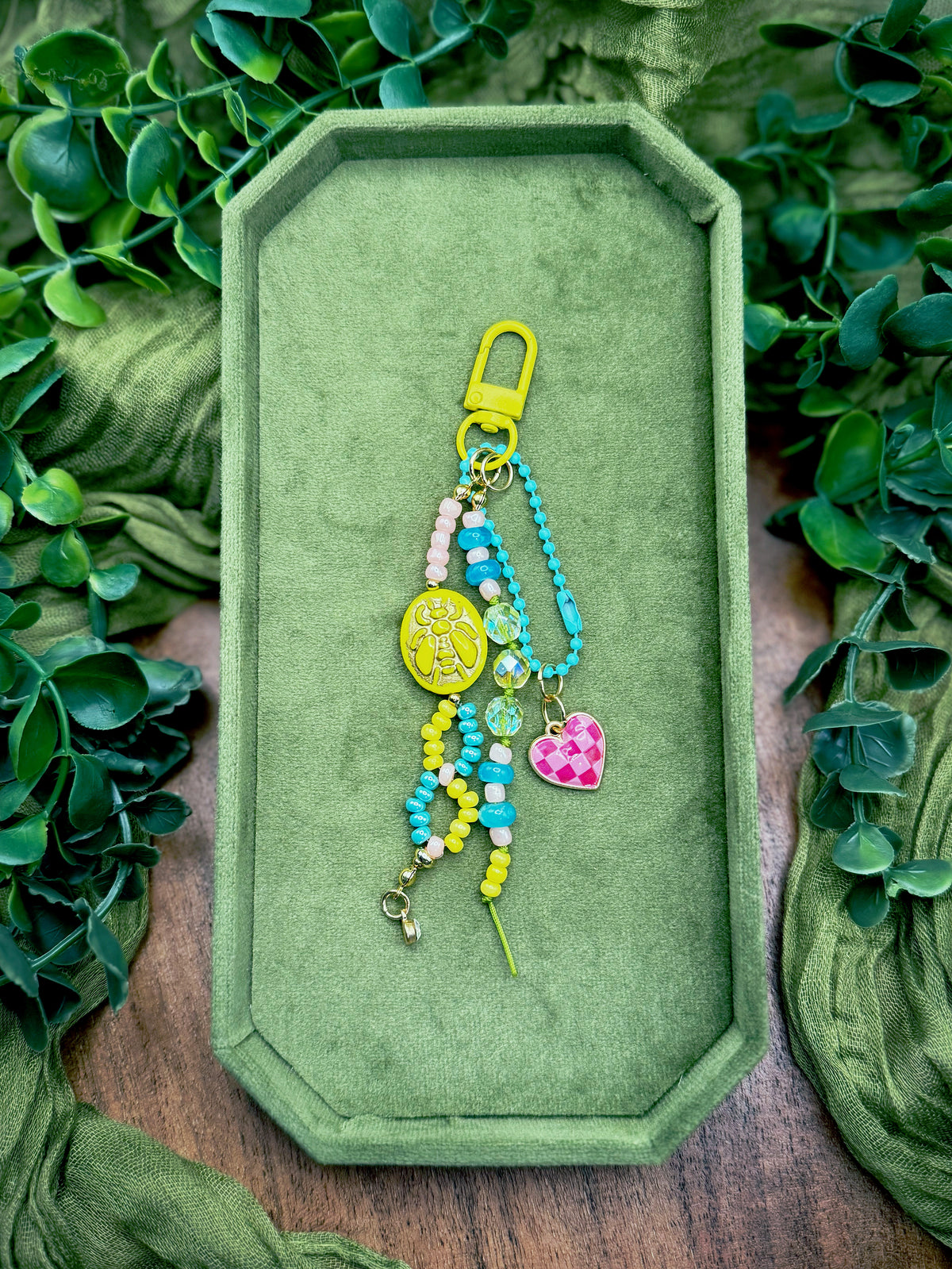 Handmade colorful beaded keychain with a yellow clasp, turquoise beads, a yellow oval charm, and a pink checkered heart charm, displayed on a green velvet tray surrounded by green leaves.