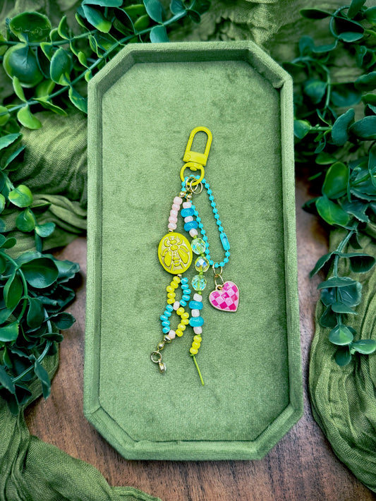 Handmade colorful beaded keychain with a yellow clasp, turquoise beads, a yellow oval charm, and a pink checkered heart charm, displayed on a green velvet tray surrounded by green leaves.