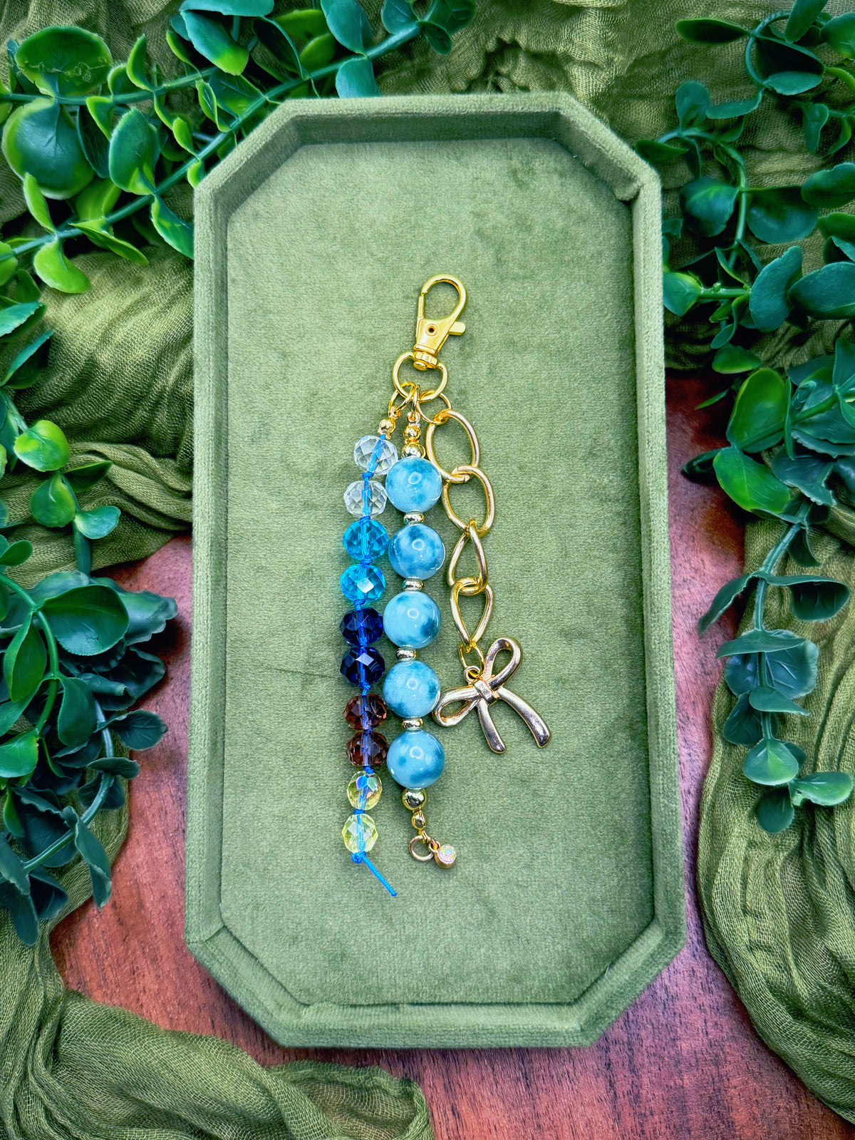 Handmade beaded charm keychain with light blue and dark blue beads, a gold clasp, and a silver bow charm, displayed on an olive green velvet tray surrounded by green leaves.