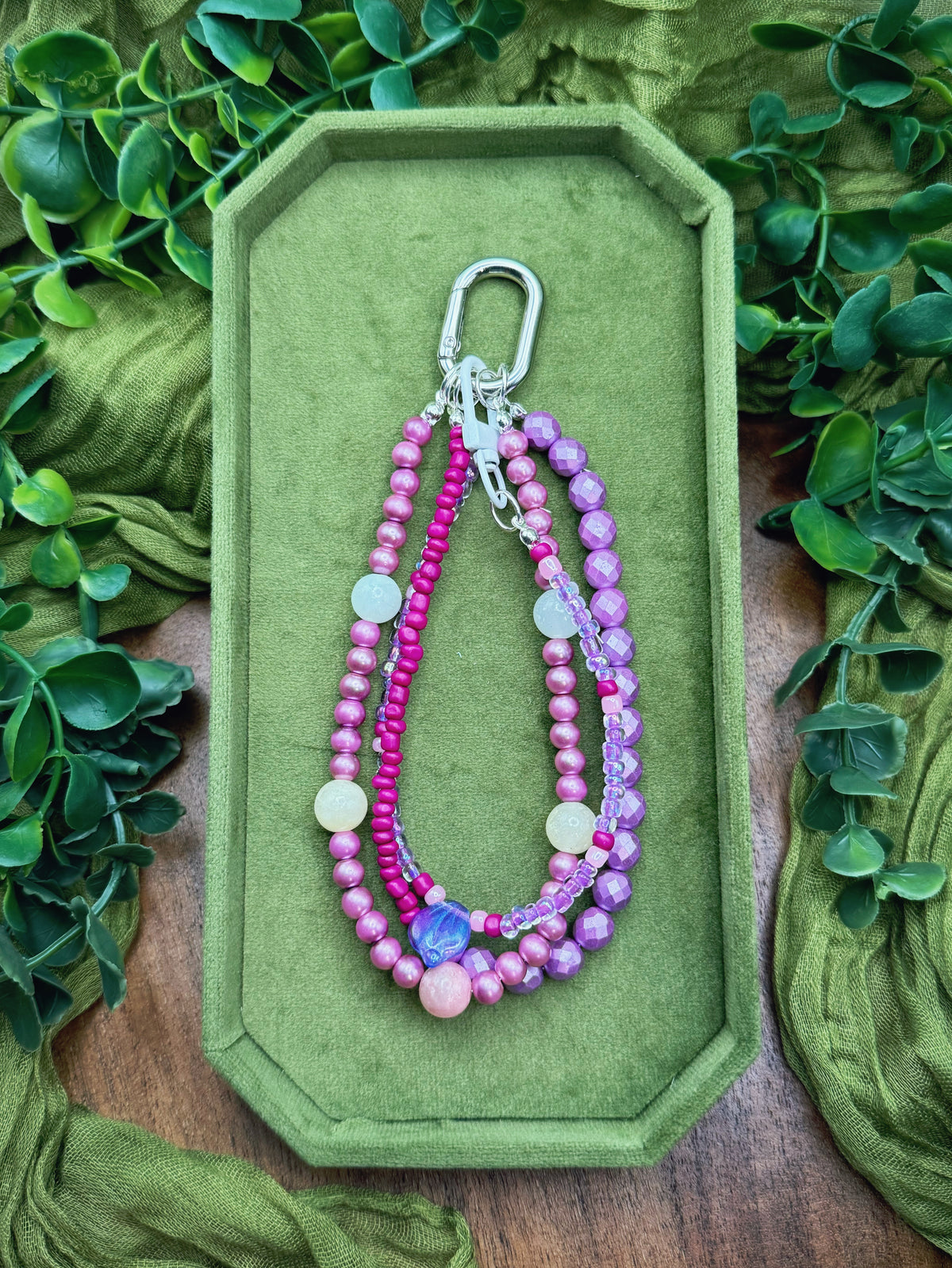 Handmade beaded keychain or bag charm with pink, purple, and white beads, a silver carabiner, and a blue bead accent, displayed on an olive green velvet tray surrounded by green leaves and fabric.