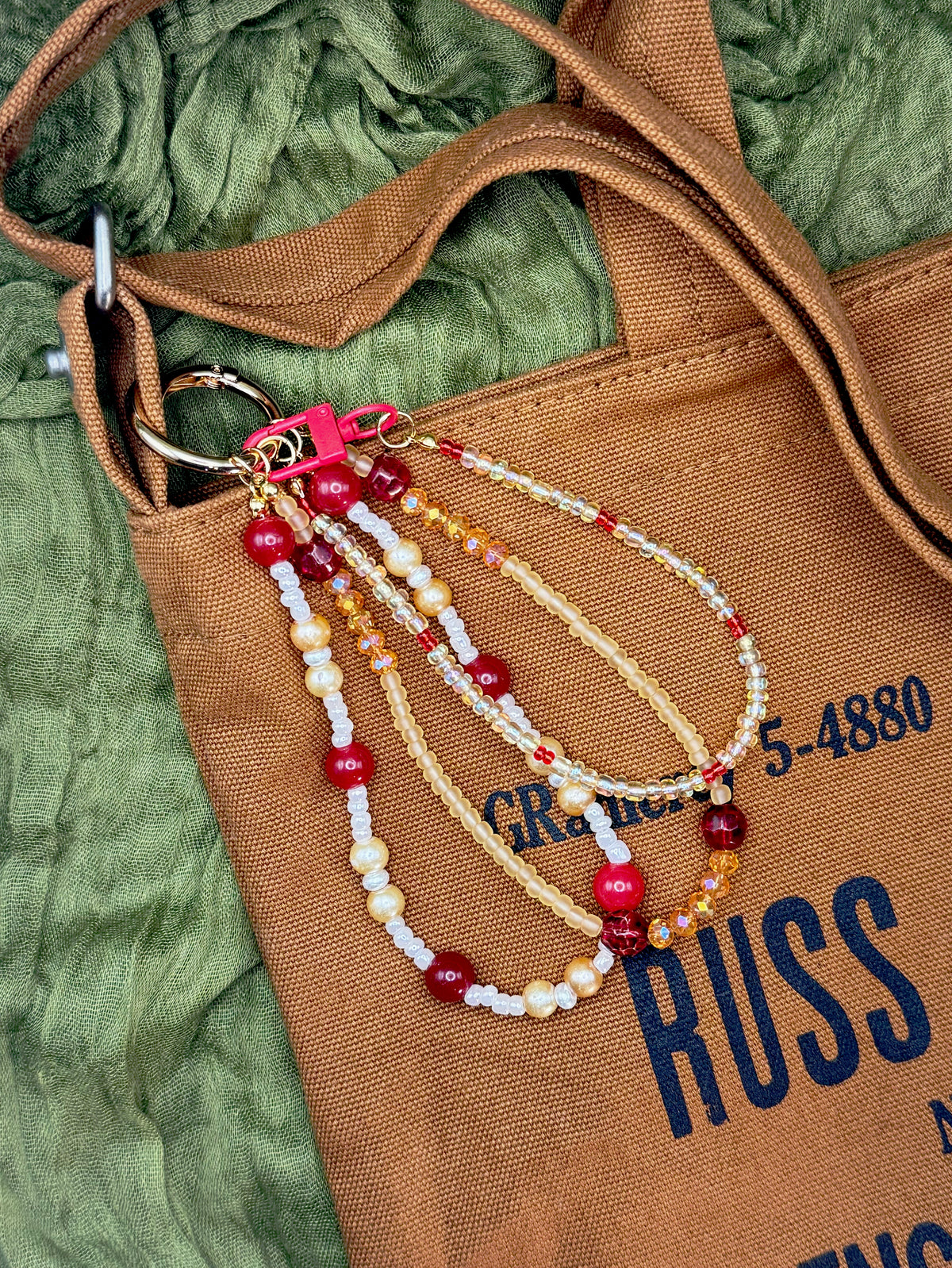 Handmade bag charm that includes cream, red, and amber beads with a pink clasp and gold-tone rings, set against a textured olive green fabric background.