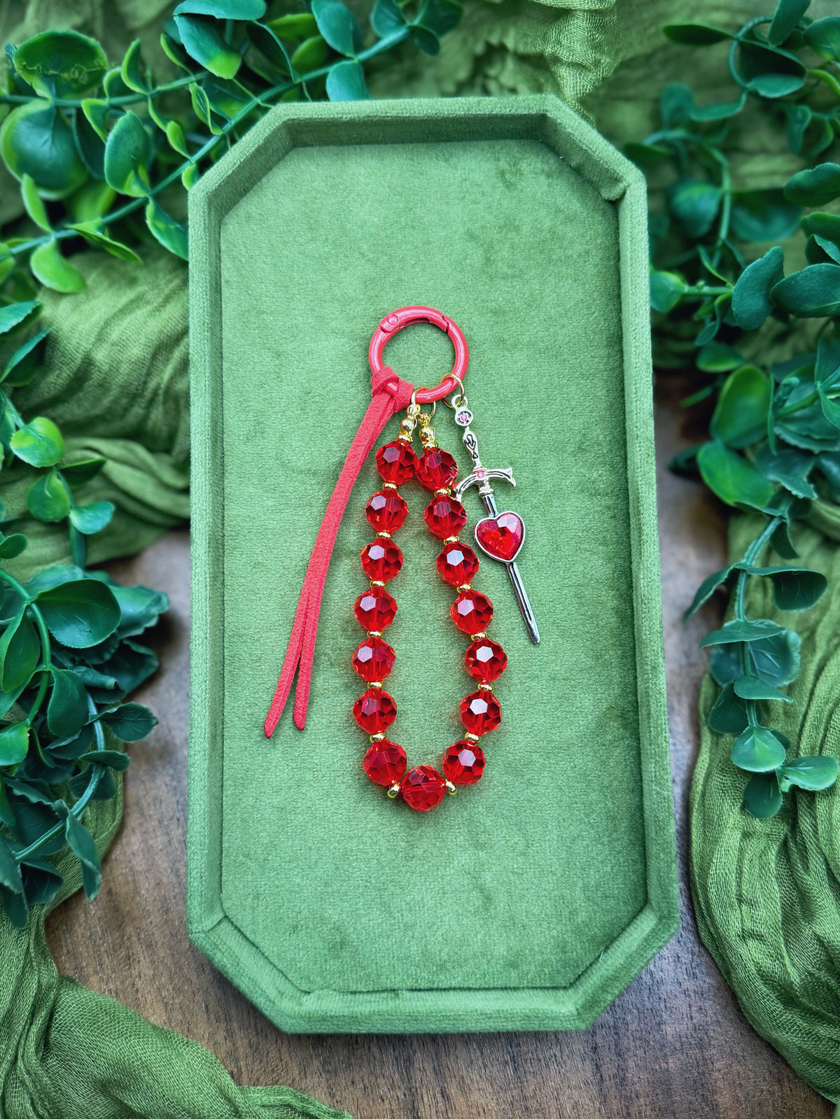 Handmade red beaded keychain with a red cord, a small silver sword charm, and a red heart pendant, displayed on a green velvet box surrounded by green leaves.