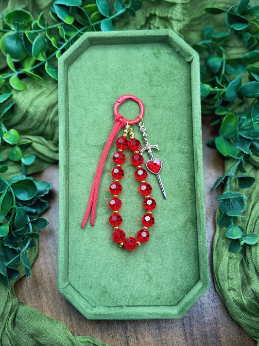 Handmade red beaded keychain with a red cord, a small silver sword charm, and a red heart pendant, displayed on a green velvet box surrounded by green leaves.
