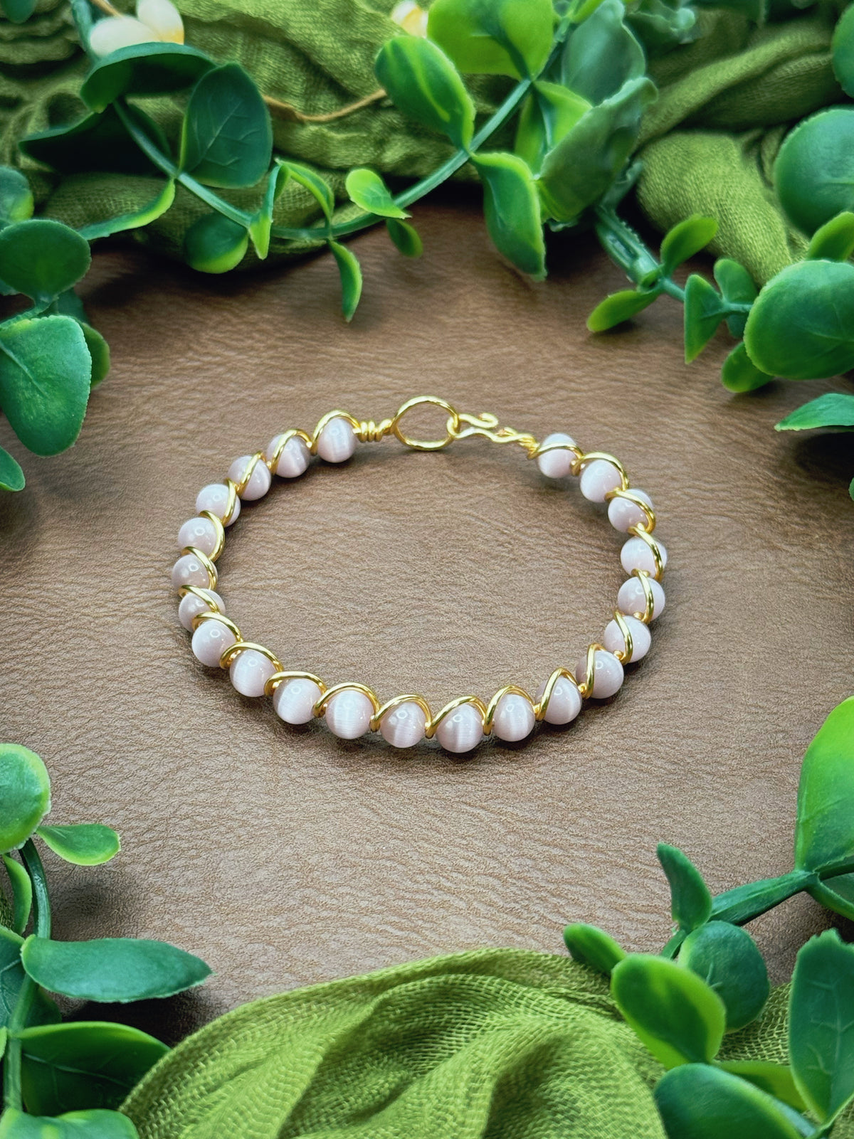 Handmade gold wire-wrapped bracelet with pale pink round beads, displayed on a brown textured surface and framed by green leaves and olive fabric.