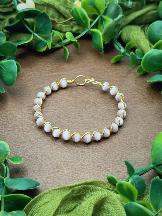 Handmade gold wire-wrapped bracelet with pale pink round beads, displayed on a brown textured surface and framed by green leaves and olive fabric.