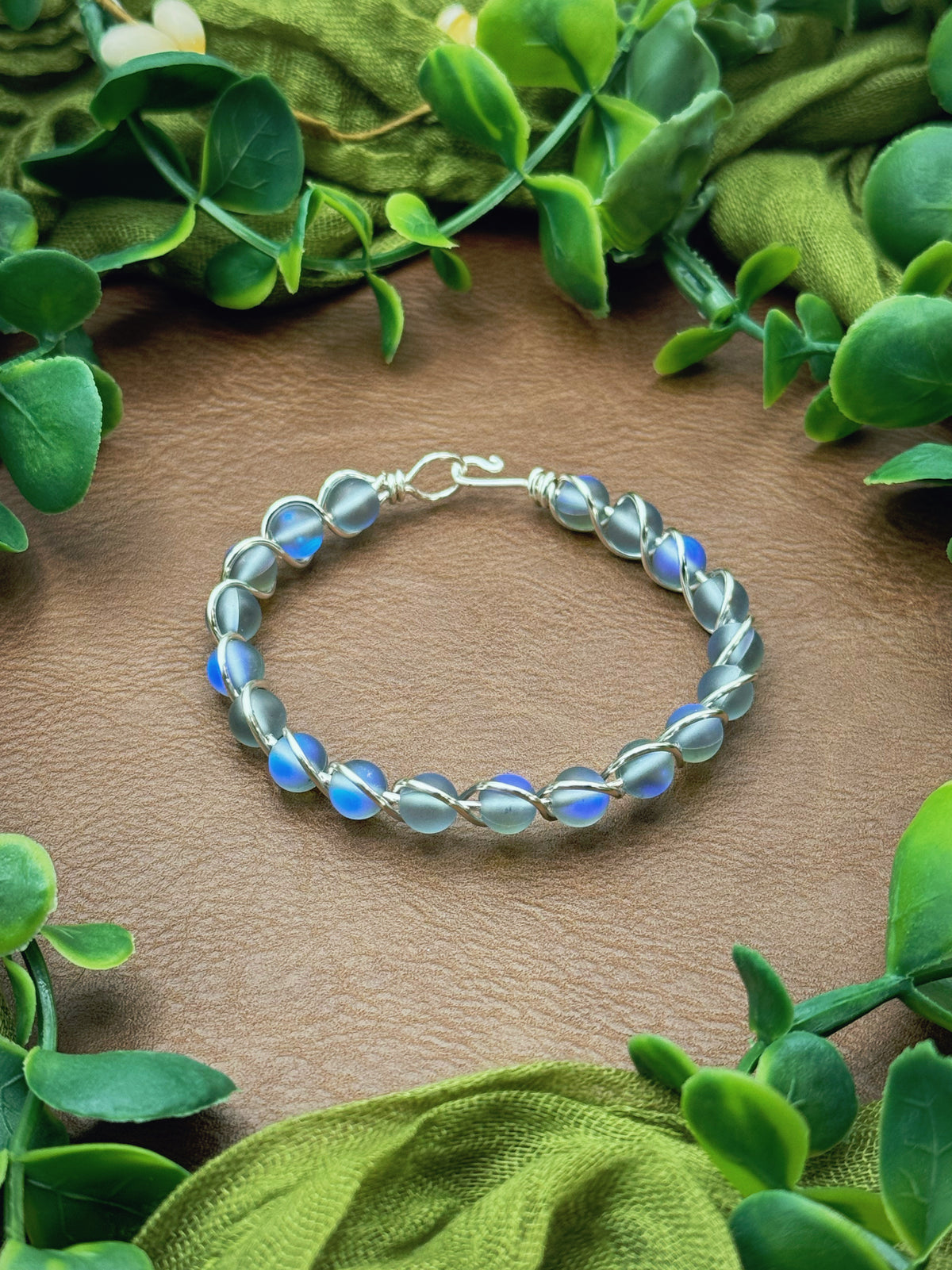 Handmade gold wire-wrapped bracelet with iridescent blue round beads, displayed on a brown textured surface and framed by green leaves and olive fabric.