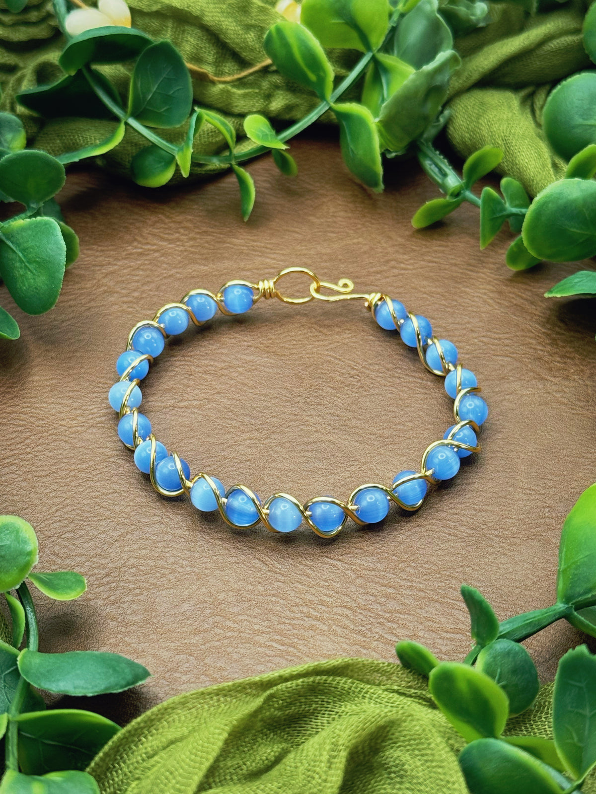 Handmade gold wire-wrapped bracelet with blue round beads, displayed on a brown textured surface and framed by green leaves and olive fabric.