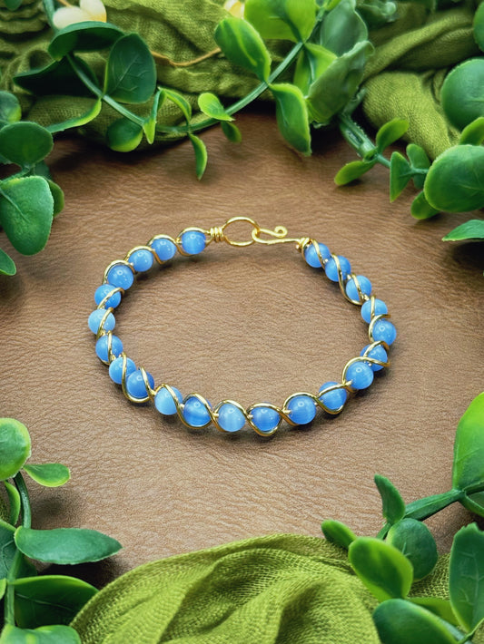Handmade gold wire-wrapped bracelet with blue round beads, displayed on a brown textured surface and framed by green leaves and olive fabric.