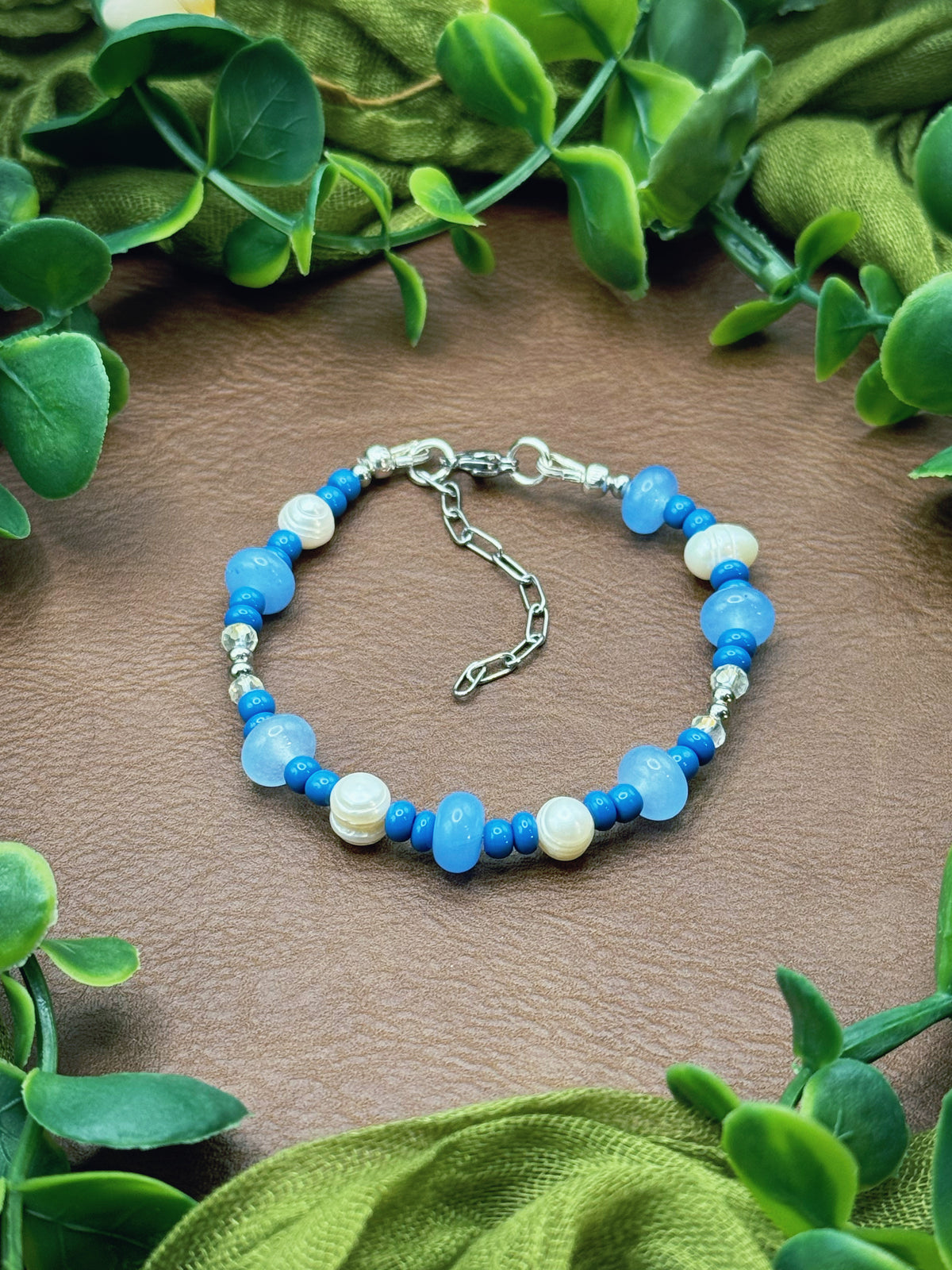 Handmade beaded bracelet with light blue and white beads, accented by silver-toned spacer beads and a silver lobster clasp, displayed on a natural surface surrounded by green leaves.