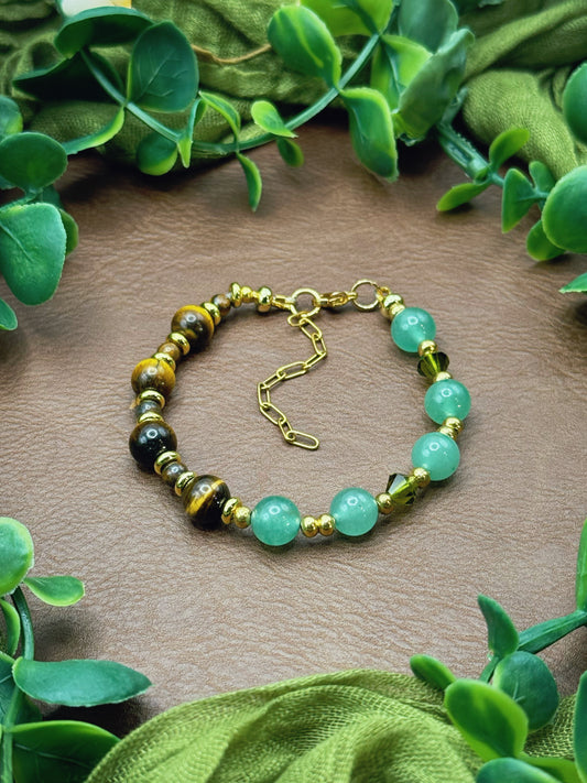 Handmade beaded bracelet with light green aventurine beads, brown tiger's eye beads, and small gold spacer beads, arranged in a circle on a natural surface with green leaves.