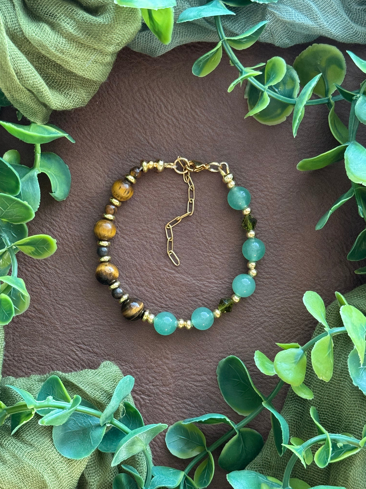 Handmade beaded bracelet with light green aventurine beads, brown tiger's eye beads, and small gold spacer beads, arranged in a circle on a natural surface with green leaves.