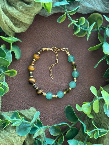 Handmade beaded bracelet with light green aventurine beads, brown tiger's eye beads, and small gold spacer beads, arranged in a circle on a natural surface with green leaves.