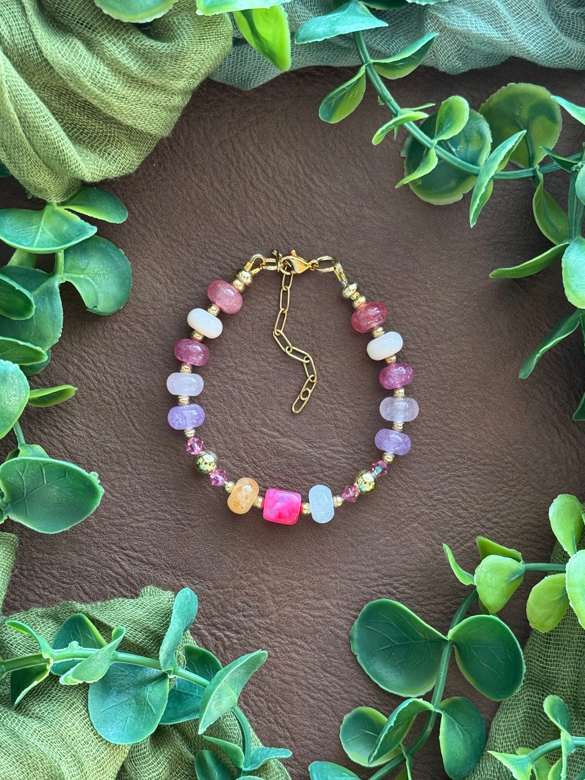 Handmade beaded bracelet with multicolored gemstone beads and gold hardware, arranged on a natural surface with green leaves.