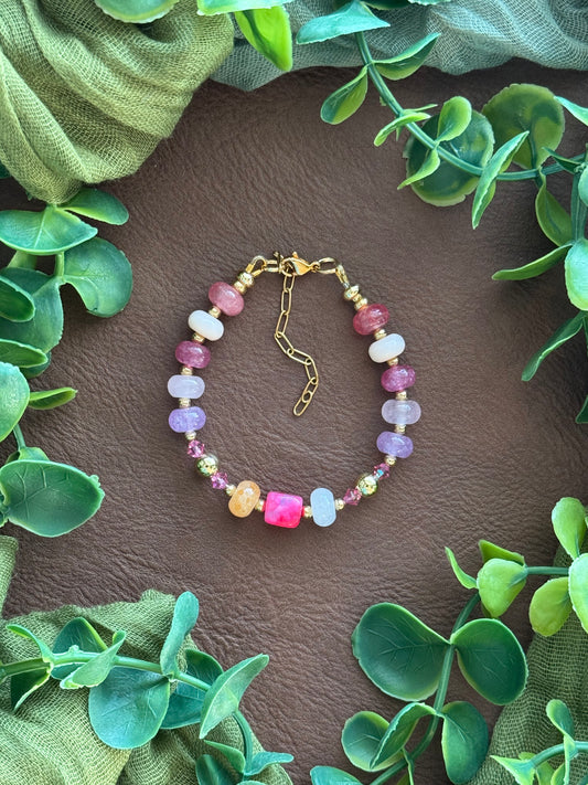 Handmade beaded bracelet with multicolored gemstone beads and gold hardware, arranged on a natural surface with green leaves.