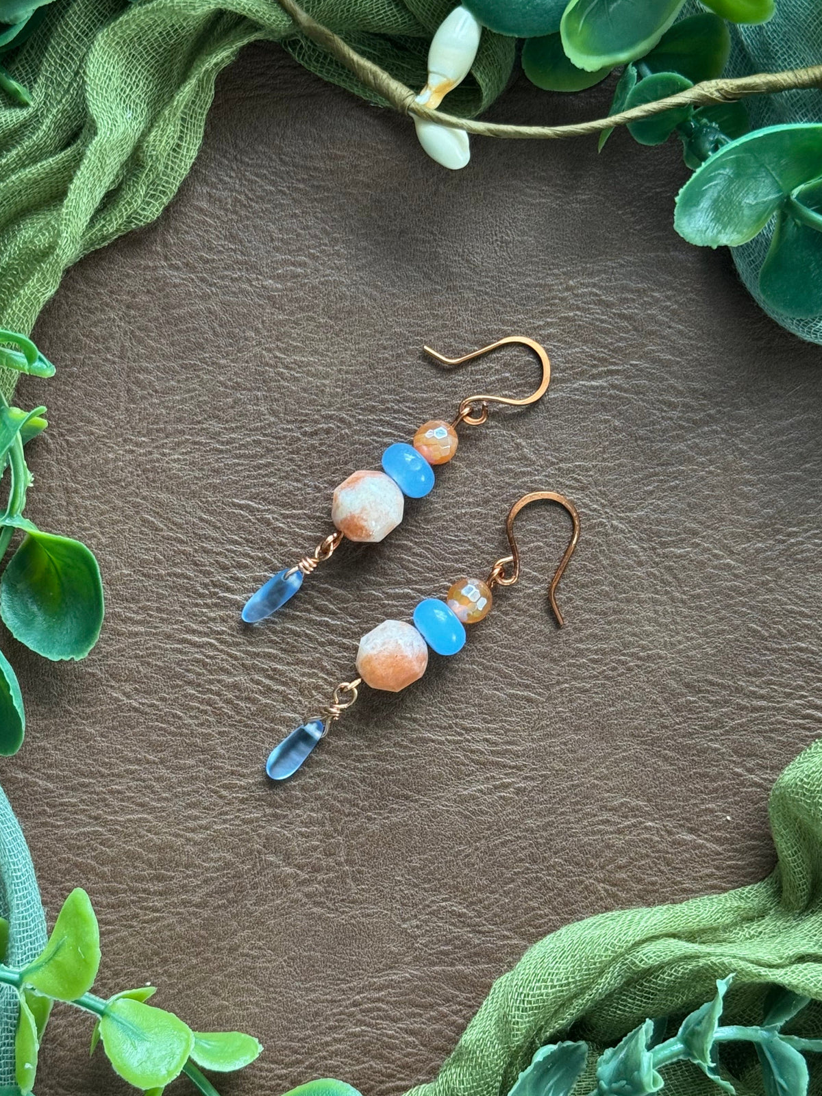 Handmade earrings with sunstone beads and gemstone beads and copper wire on a natural surface surrounded by green leaves.