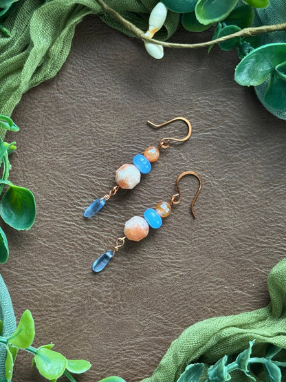 Handmade earrings with sunstone beads and gemstone beads and copper wire on a natural surface surrounded by green leaves.