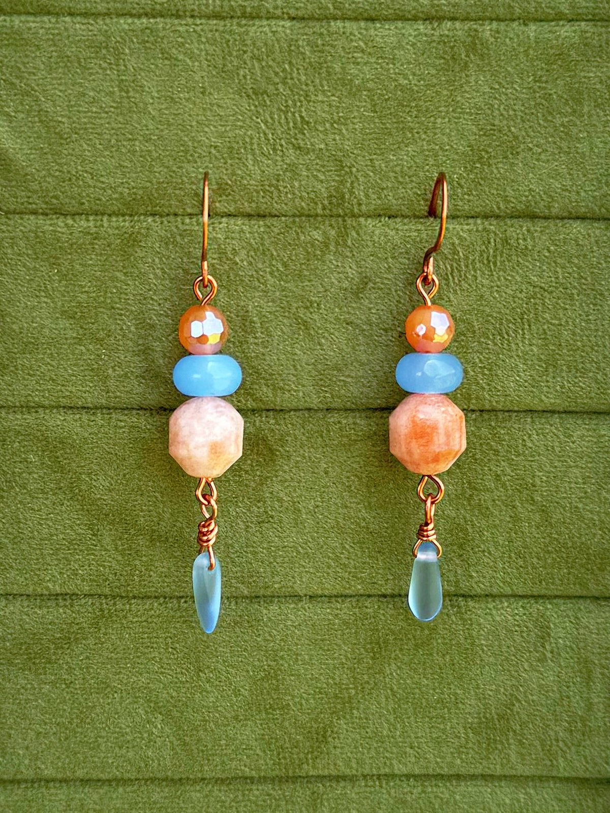 Handmade dangle earrings with sunstone, light blue, and cosmic agate faceted beads and copper wire hooks, displayed on a textured olive green background.