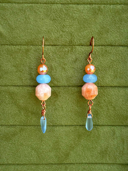 Handmade dangle earrings with sunstone, light blue, and cosmic agate faceted beads and copper wire hooks, displayed on a textured olive green background.