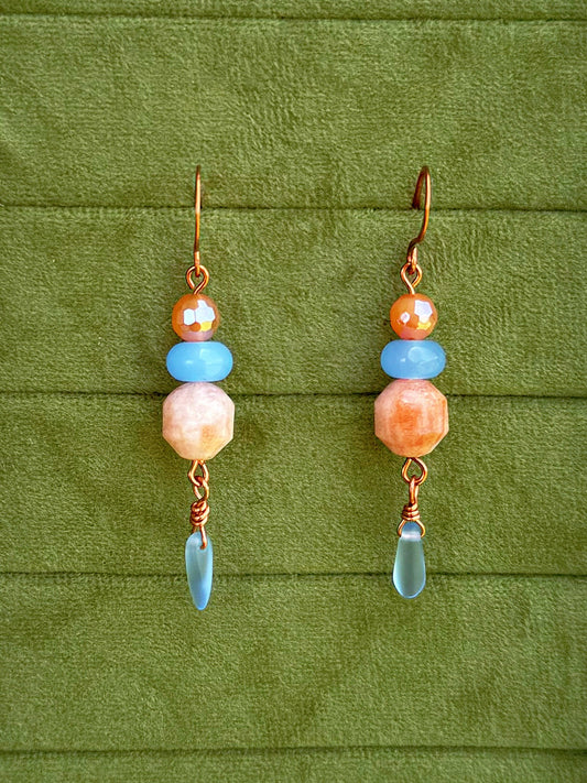 Handmade dangle earrings with sunstone, light blue, and cosmic agate faceted beads and copper wire hooks, displayed on a textured olive green background.