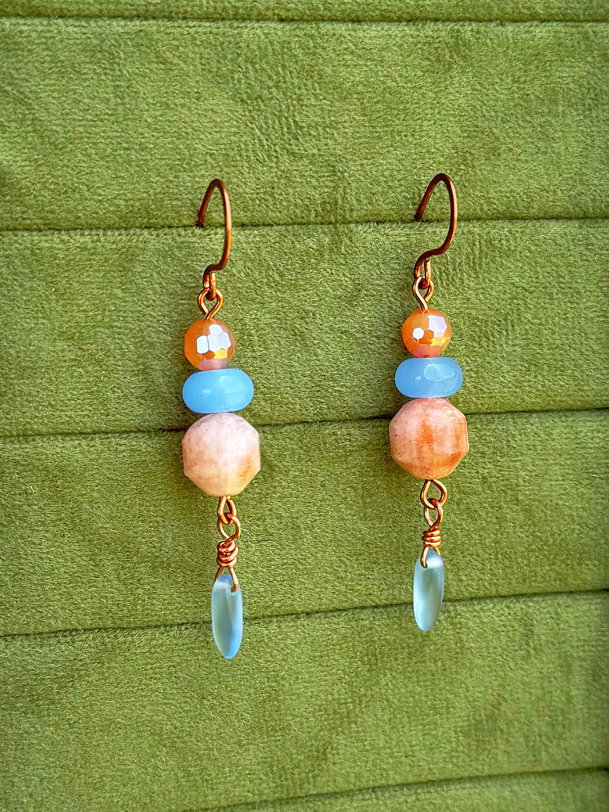 Handmade dangle earrings with sunstone, light blue, and cosmic agate faceted beads and copper wire hooks, displayed on a textured olive green background.
