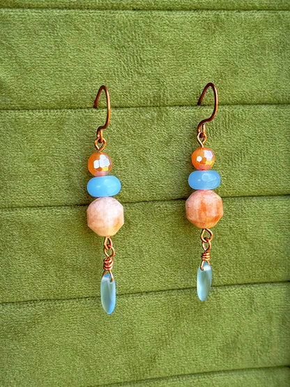 Handmade dangle earrings with sunstone, light blue, and cosmic agate faceted beads and copper wire hooks, displayed on a textured olive green background.