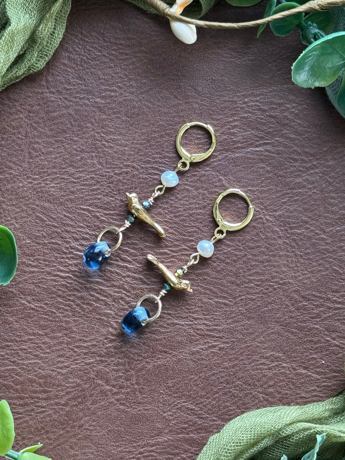 Handmade beaded earrings with gemstone and glass beads, featuring a gold bird charm on a natural background with green leaves around.