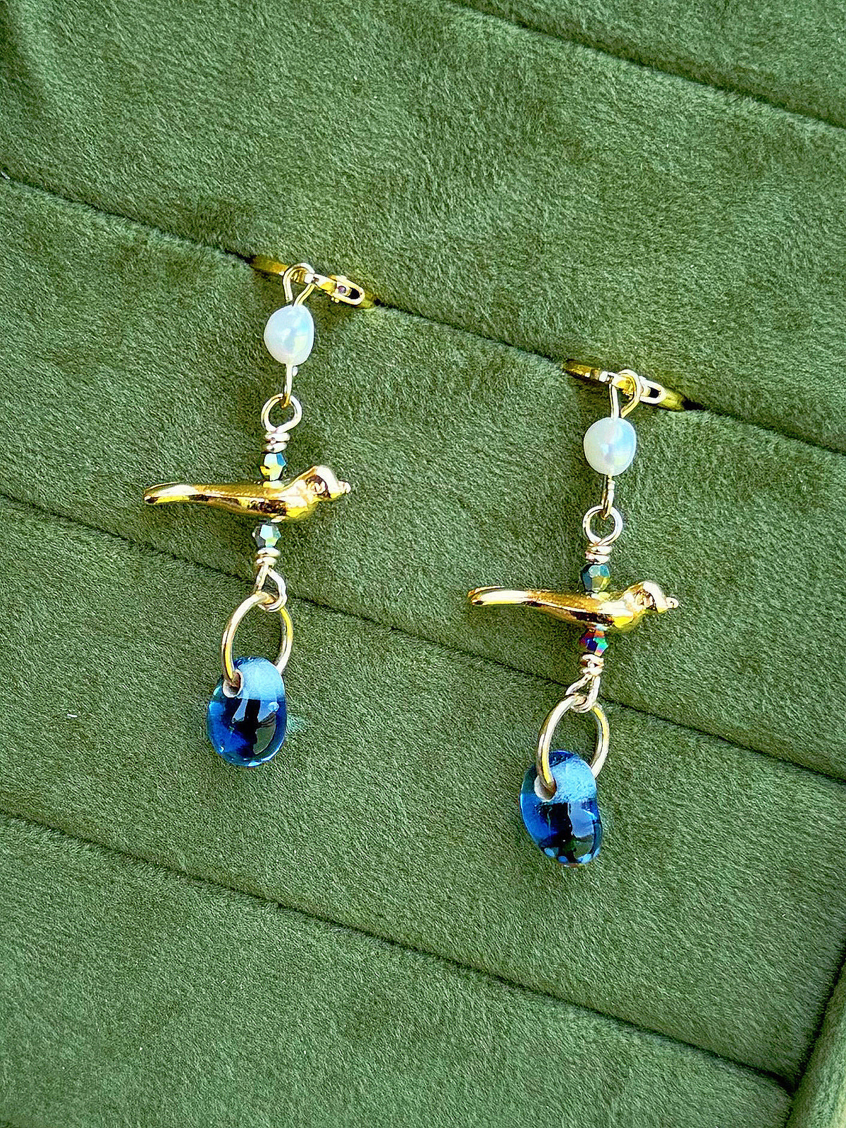 Handmade beaded earrings with gemstone and glass beads, featuring a gold bird charm displayed on a textured olive green background.