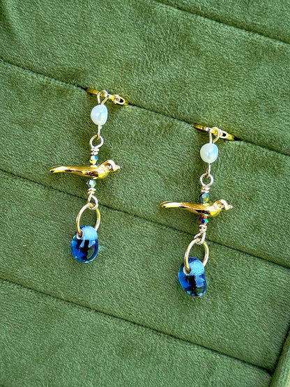 Handmade beaded earrings with gemstone and glass beads, featuring a gold bird charm displayed on a textured olive green background.