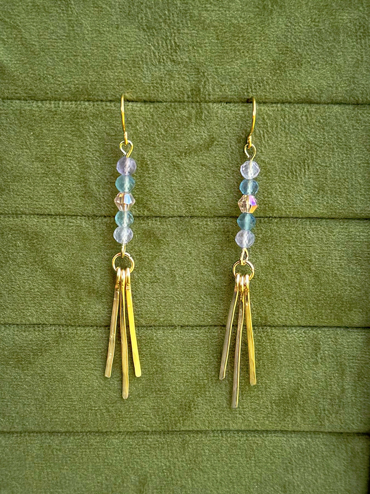 Handmade earrings with small iridescent beads and three gold-toned fringe bars, displayed on a textured olive green fabric background.