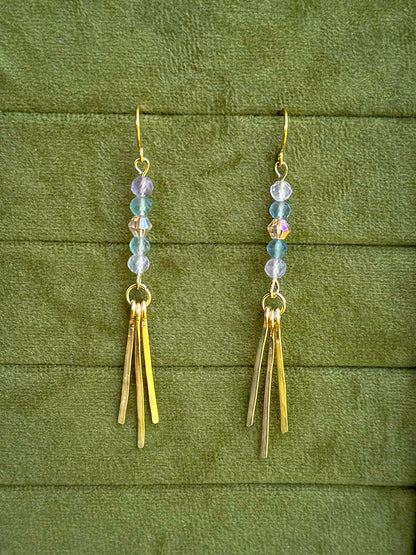 Handmade earrings with small iridescent beads and three gold-toned fringe bars, displayed on a textured olive green fabric background.
