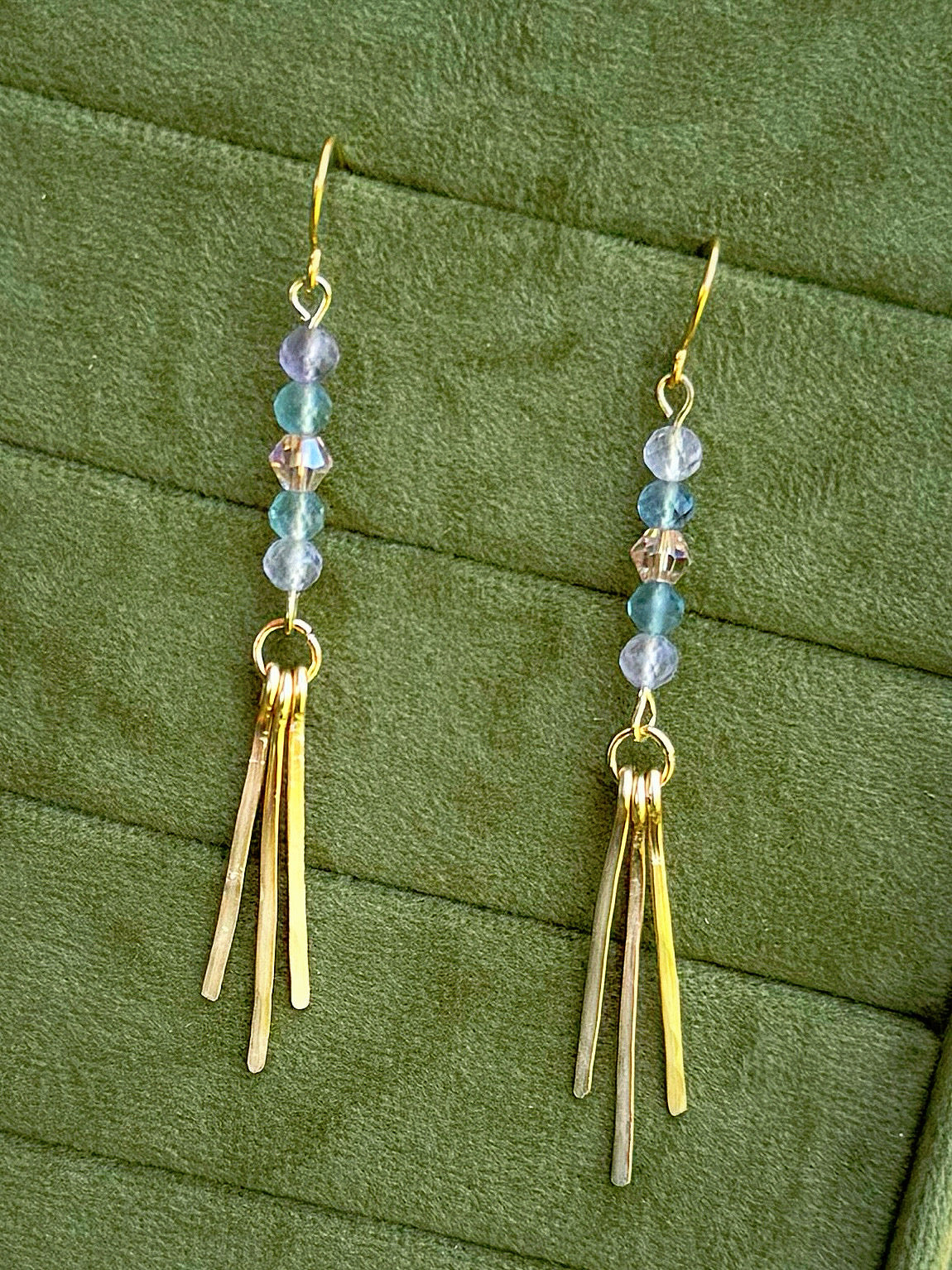 Handmade earrings with small iridescent beads and three gold-toned fringe bars, displayed on a textured olive green fabric background.
