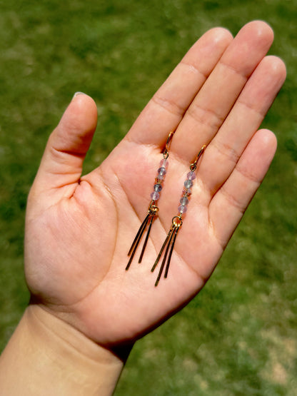 Handmade earrings with small iridescent beads and three gold-toned fringe bars on a green grass background.