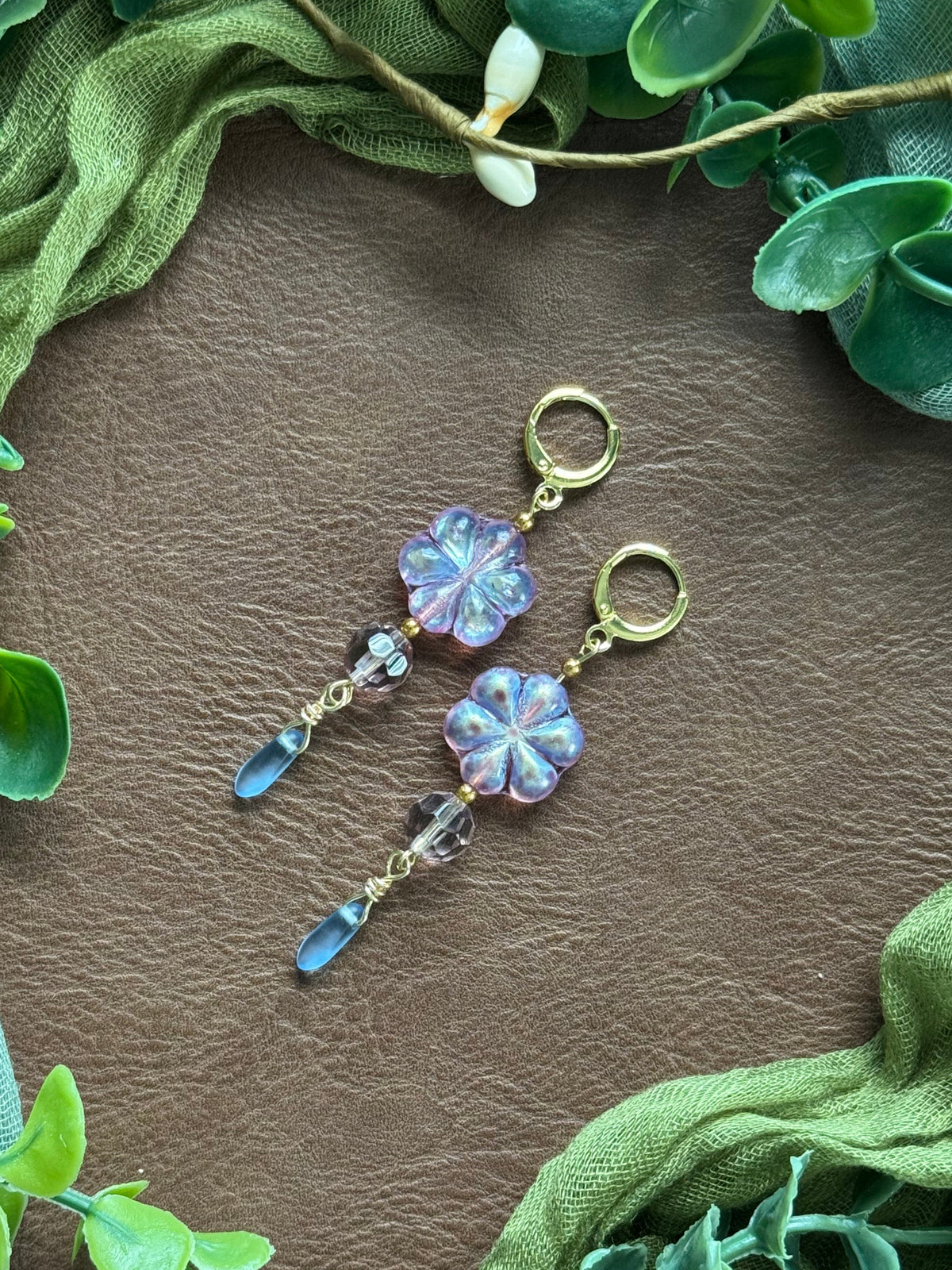 Handmade earrings with lavender-blue flower-shaped beads and clear faceted drops, arranged on a natural surface surrounded by green leaves.