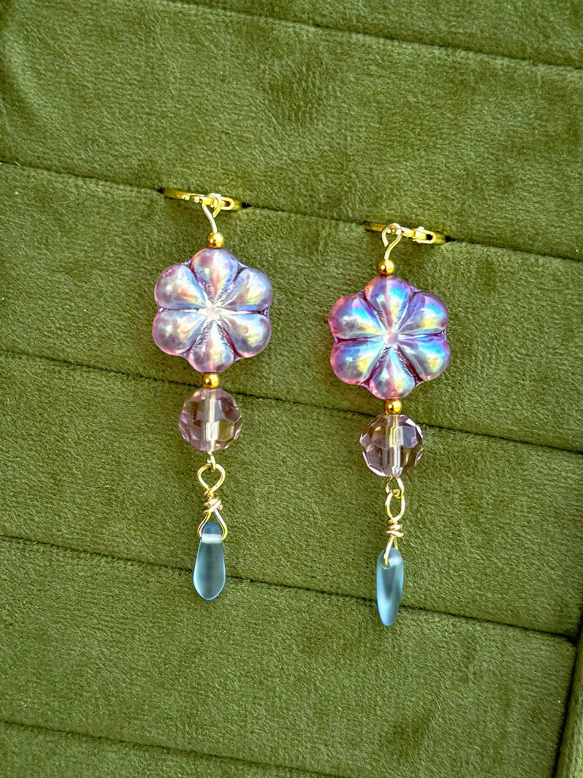 Handmade earrings with lavender-blue flower-shaped beads and clear faceted drops, arranged on an olive green velvet background.