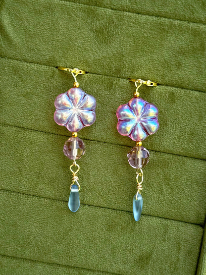 Handmade earrings with lavender-blue flower-shaped beads and clear faceted drops, arranged on an olive green velvet background.