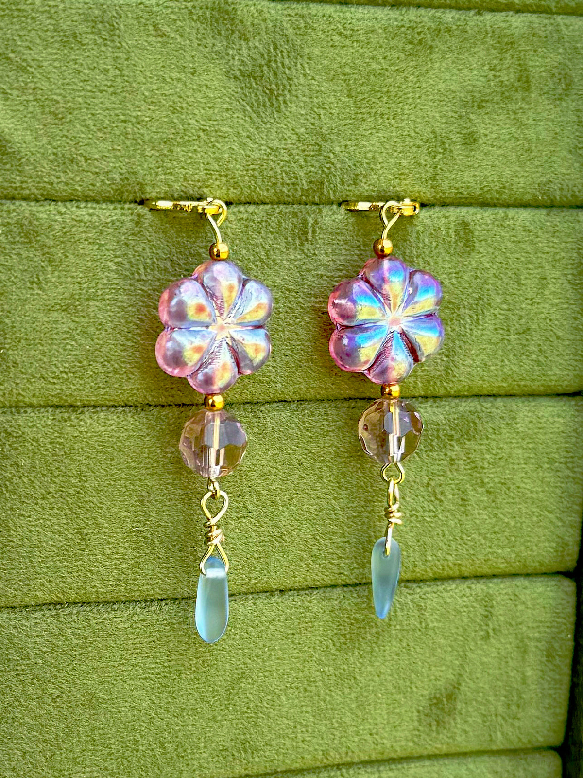 Handmade earrings with lavender-blue flower-shaped beads and clear faceted drops, arranged on an olive green velvet background.