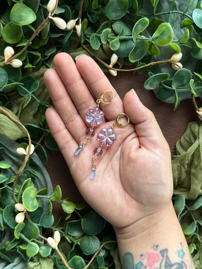 Handmade earrings with lavender-blue flower-shaped beads and clear faceted drops, arranged on a natural surface surrounded by green leaves.