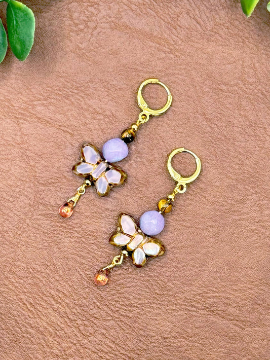 Handmade earrings with butterfly charms, light purple beads, and small amber drop beads, displayed on a textured brown surface with green leaves around the edges.
