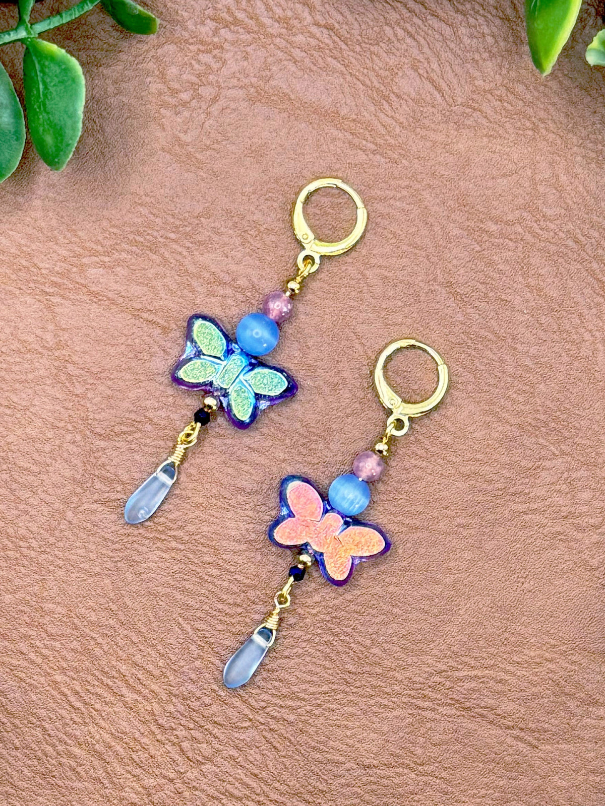 Handmade earrings featuring butterfly charms in blue and pink, with gold-tone hoops and clear crystal drops, displayed on a textured brown leather background with green leaves.